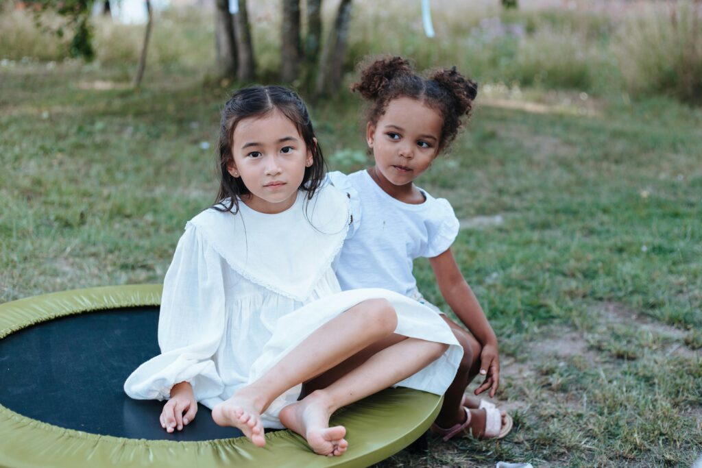 Two young girls in white dresses sitting on a trampoline in an outdoor setting.